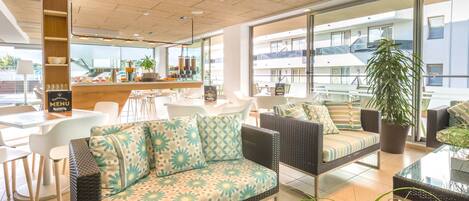 A modern hotel lobby with a sofa, chairs, a bar area, and large windows offering views of the building.