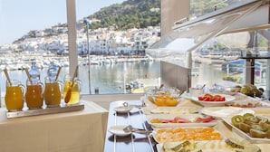 Een buffet tafel met verschillende soorten fruit en drankjes, met uitzicht op een jachthaven en een heuvelstad.