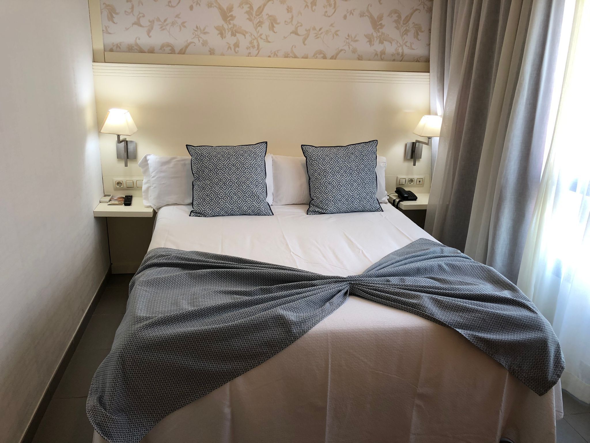 Een hotelkamer met een netjes opgemaakt bed, twee gestreepte kussens, een bedlampje, een telefoon en een gordijn.