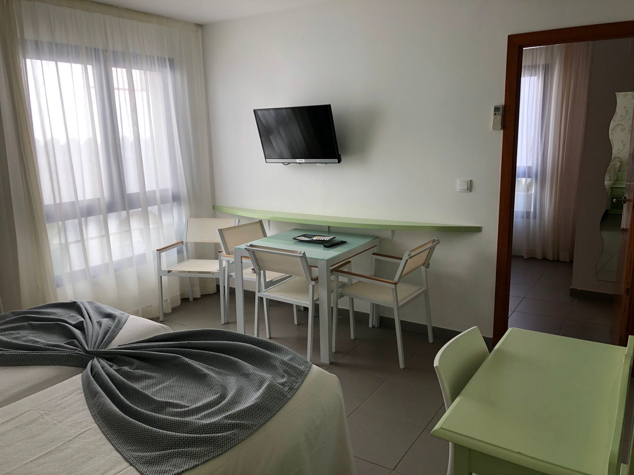 A hotel room with a bed, a desk with chairs, a television mounted on the wall, and a window with sheer curtains.