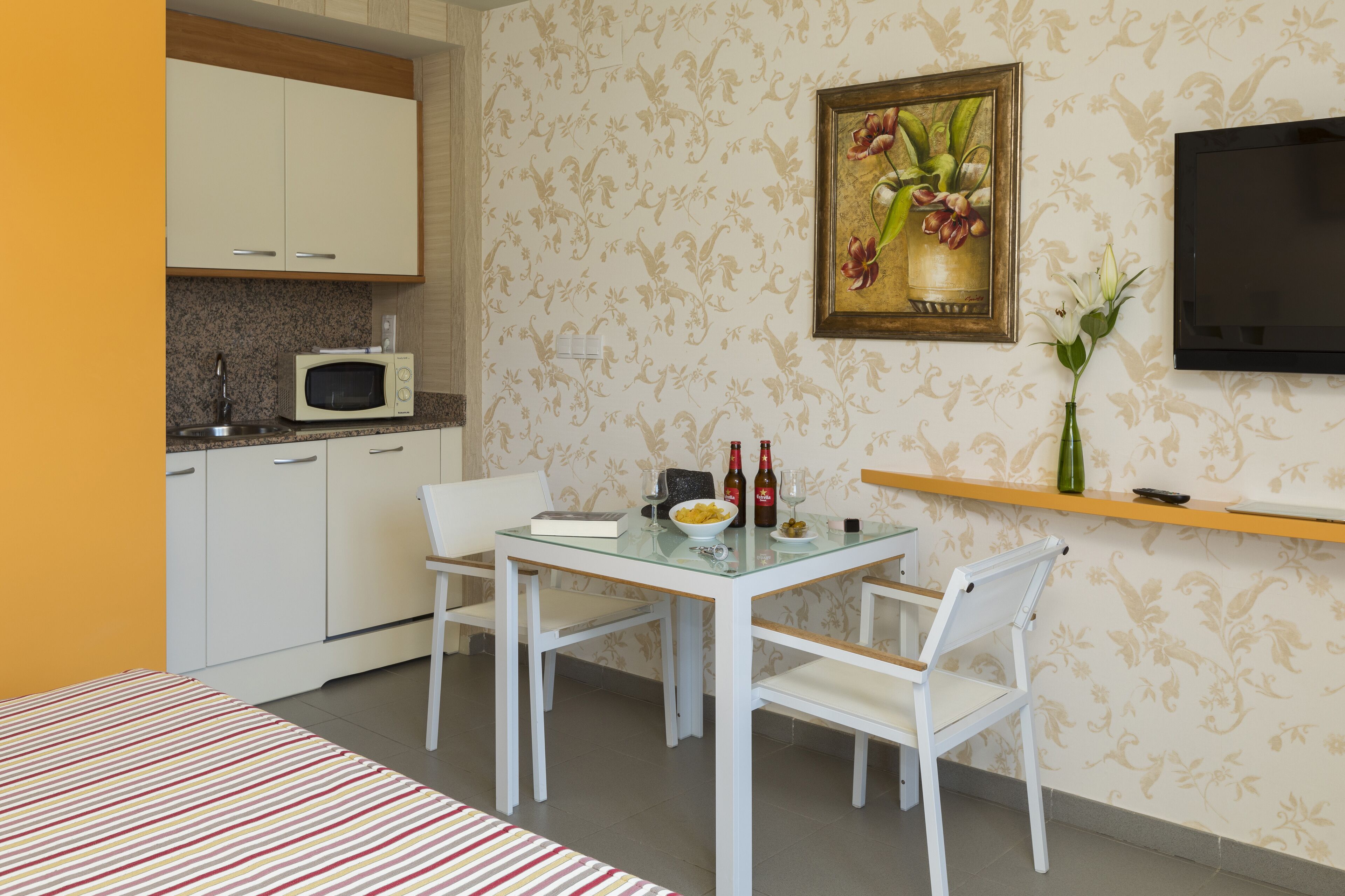 Una habitación compacta con kitchenette, una mesa de comedor con sillas, una cama y un televisor montado en la pared.
