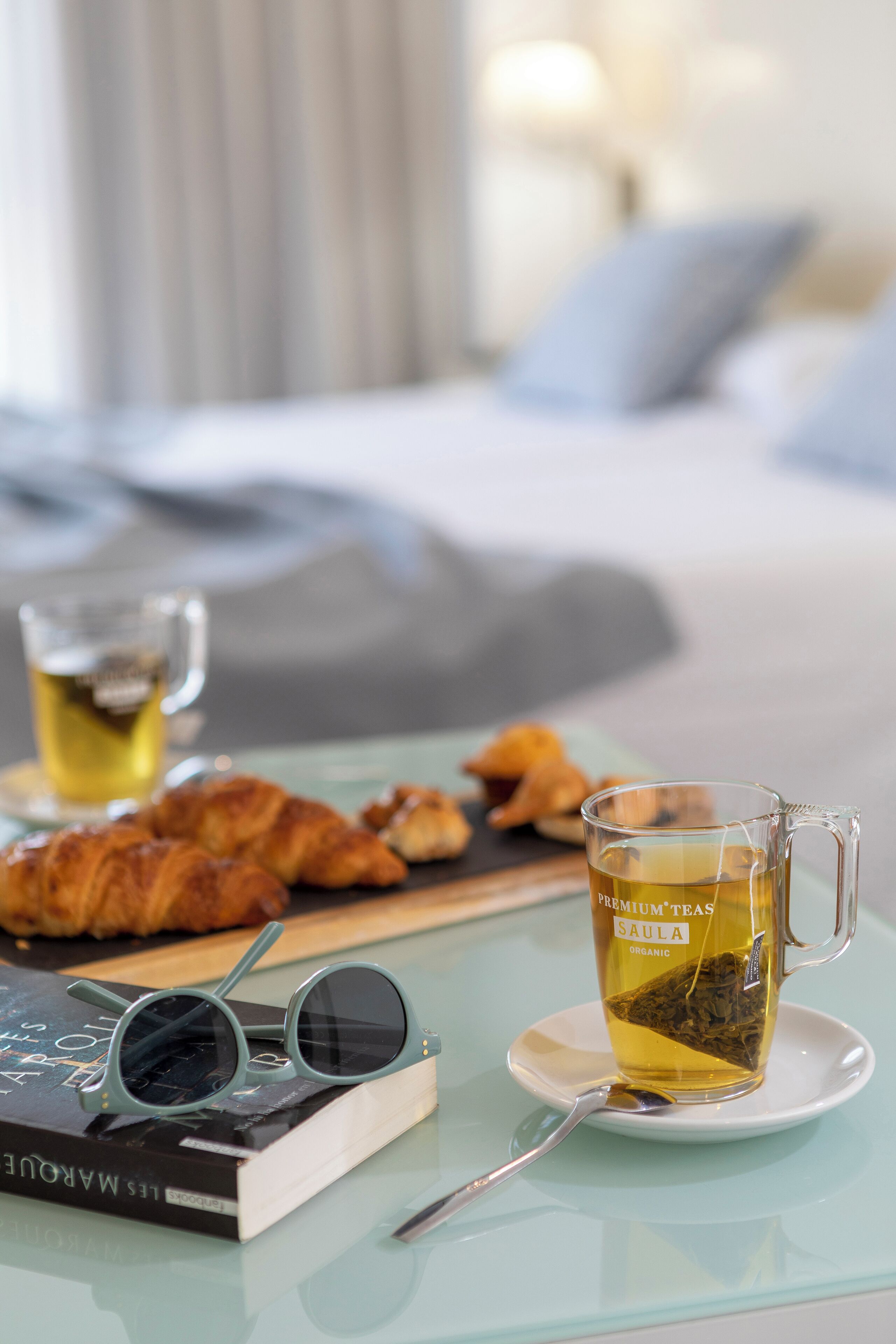 Una taza de vidrio con té, un libro y croissants sobre una mesa.