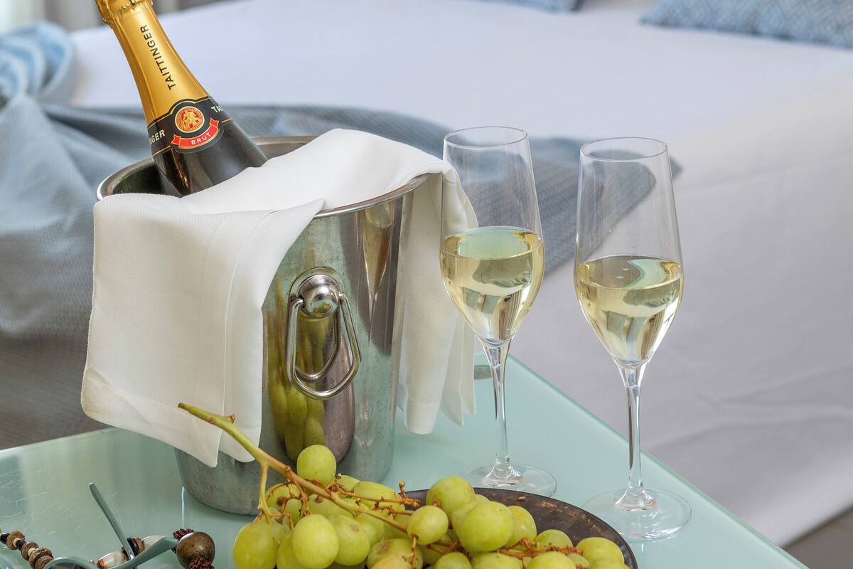 A bed with white linens and blue pillows, a bedside table with a champagne bottle, glasses, and a plate of grapes.