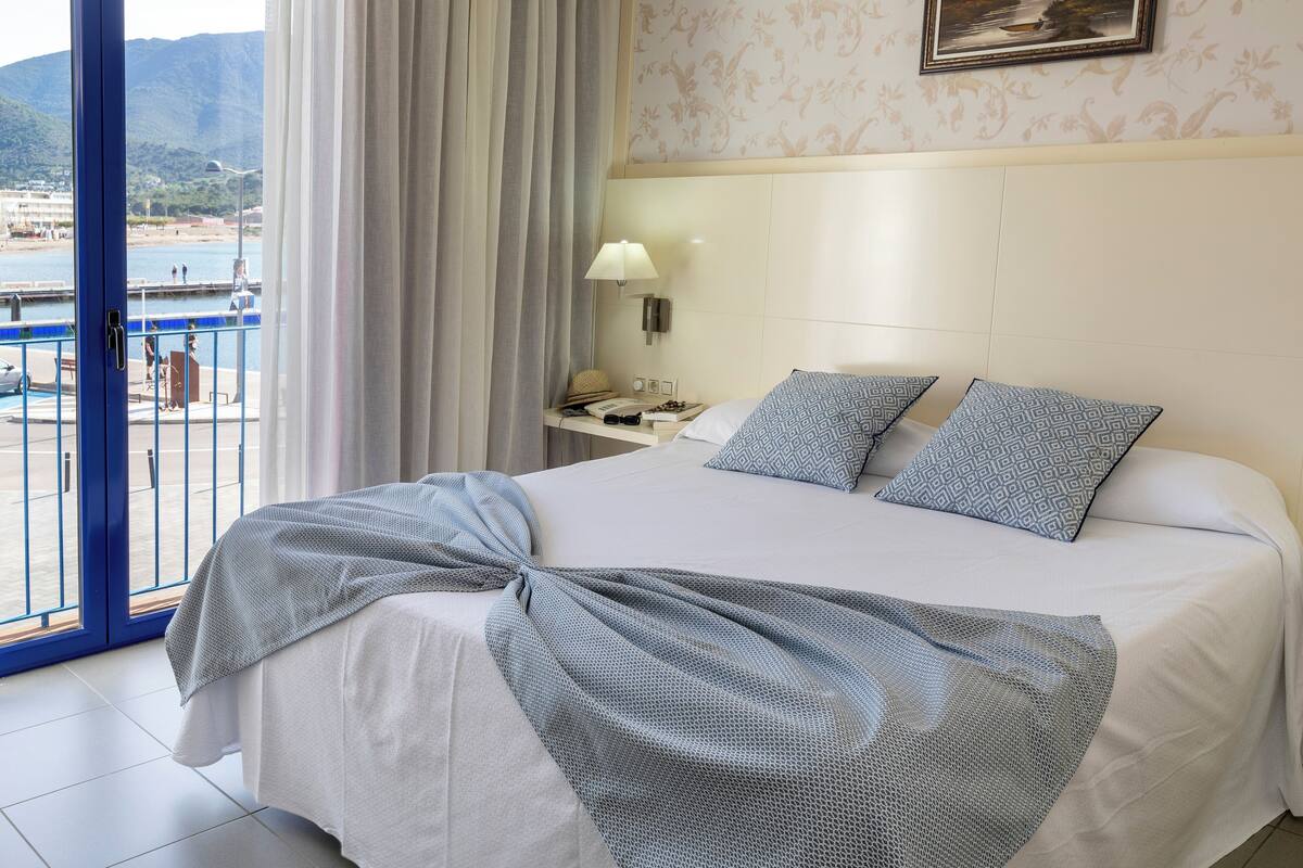 A hotel room with a bed, a nightstand, a lamp, and a view of a body of water through a balcony door.