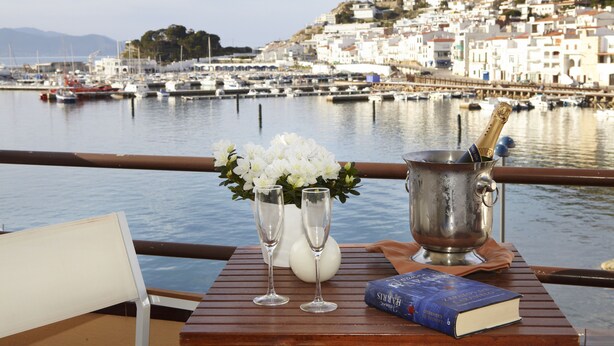 Een tafel met een boek, twee champagneglazen en een bloemstuk, met uitzicht op een waterkant met boten en gebouwen op de achtergrond.