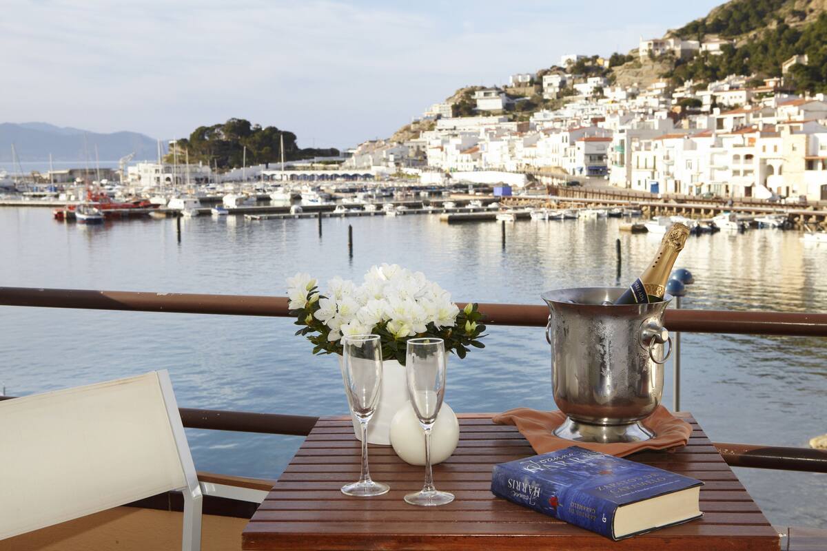 A table with a book, two champagne flutes, and a flower arrangement by a waterfront with boats and buildings in the background.