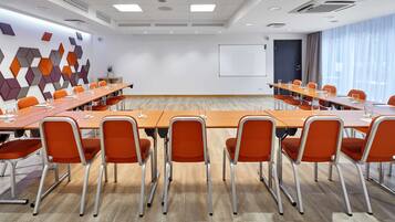 Un salón de conferencias con varias mesas y sillas naranja, paredes blancas y un diseño geométrico en la pared.