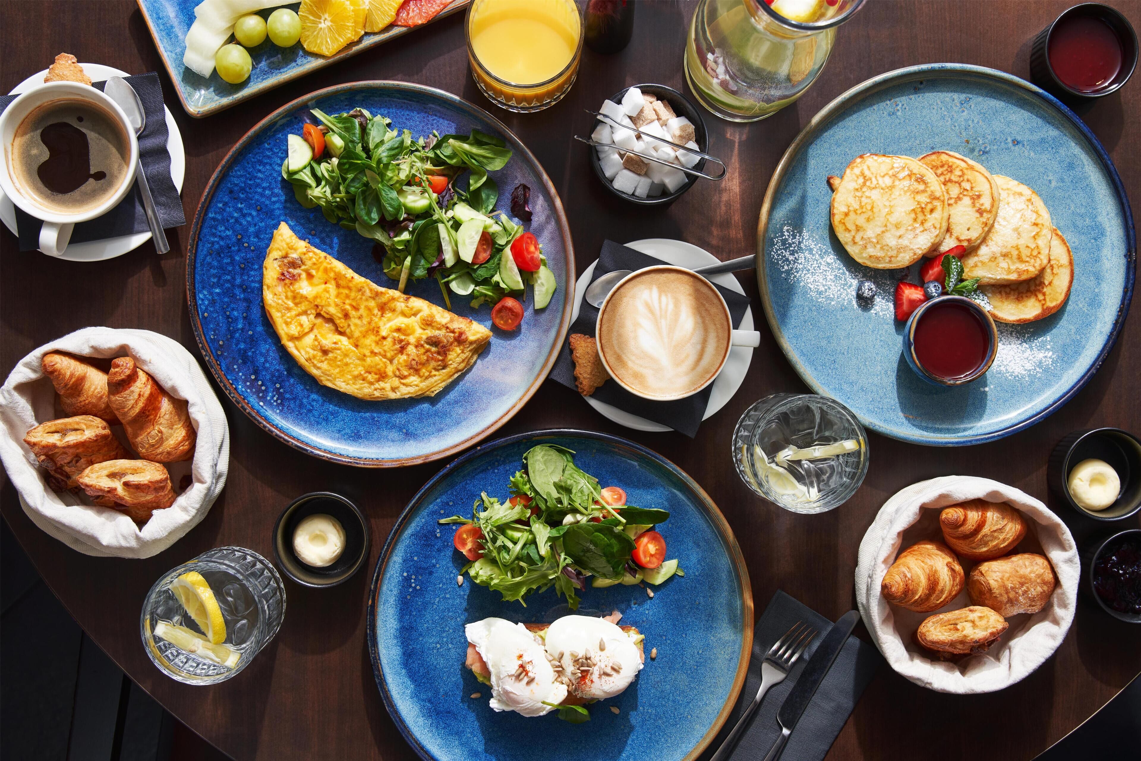 Una escena de desayuno con platos de comida, incluyendo huevos, tostadas y una ensalada, junto con bebidas y pasteles.