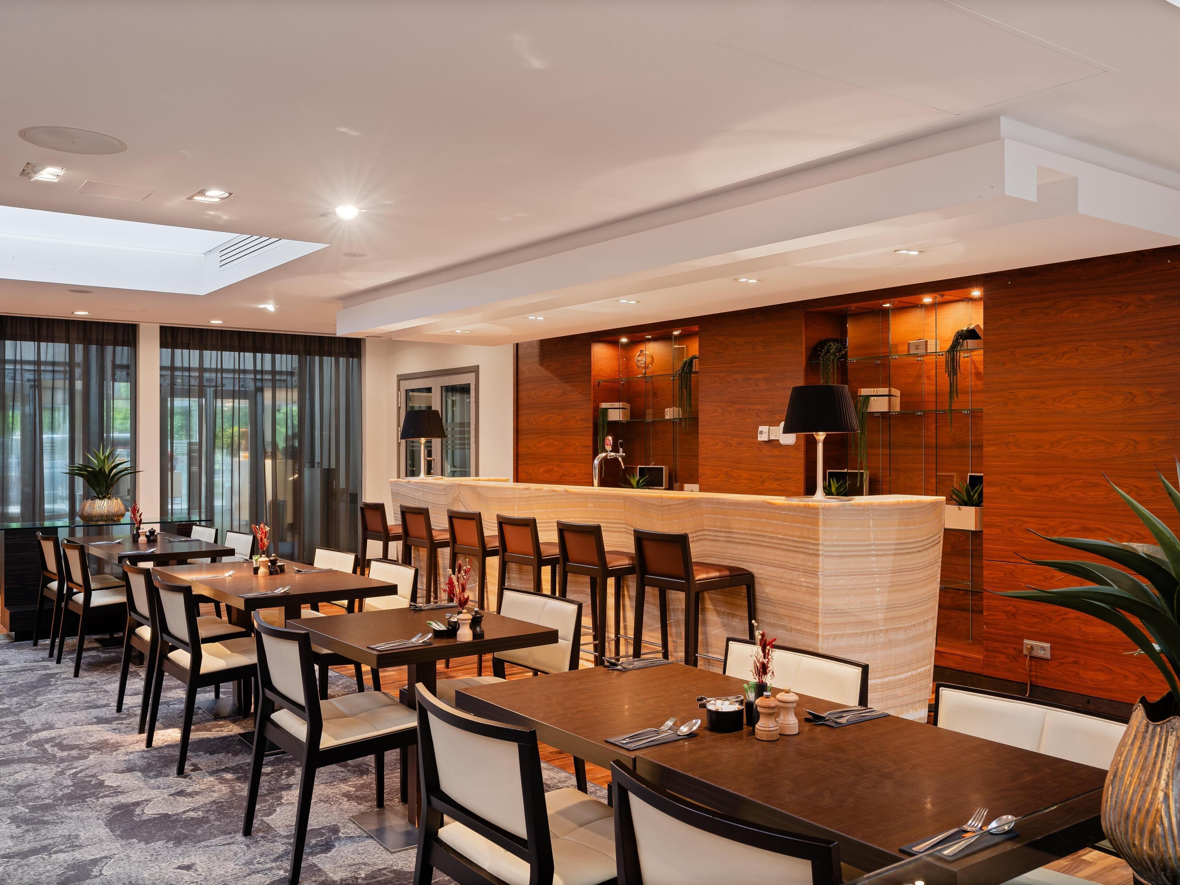 A modern hotel dining area with multiple tables and chairs, a central bar, and a view of the outdoors.