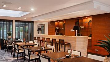 A modern hotel dining area with multiple tables and chairs, a central bar, and a view of the outdoors.