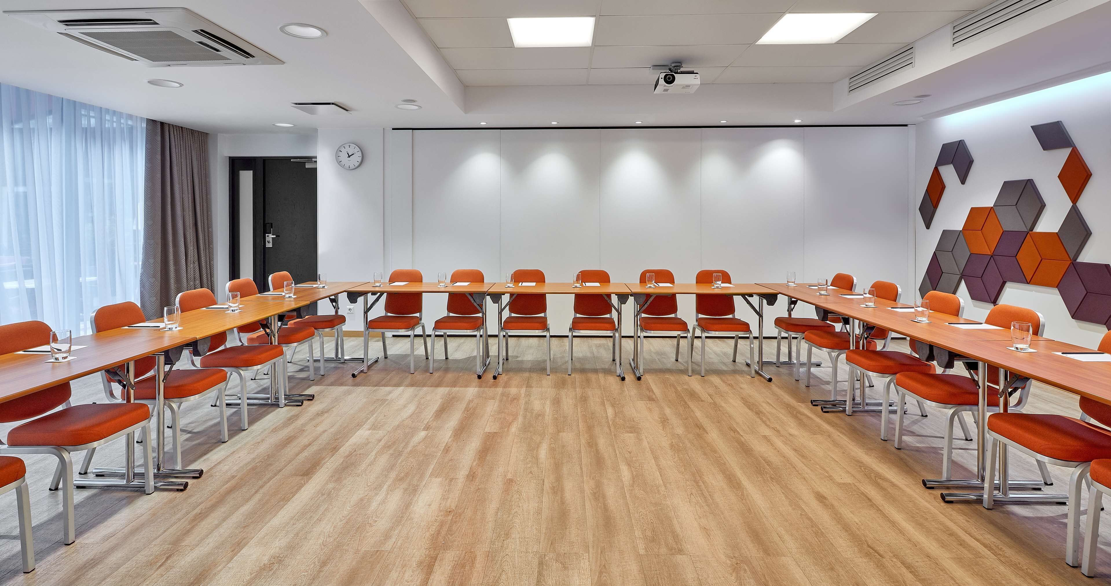 A conference room with a long table, orange chairs, a clock, and a projector on the ceiling.