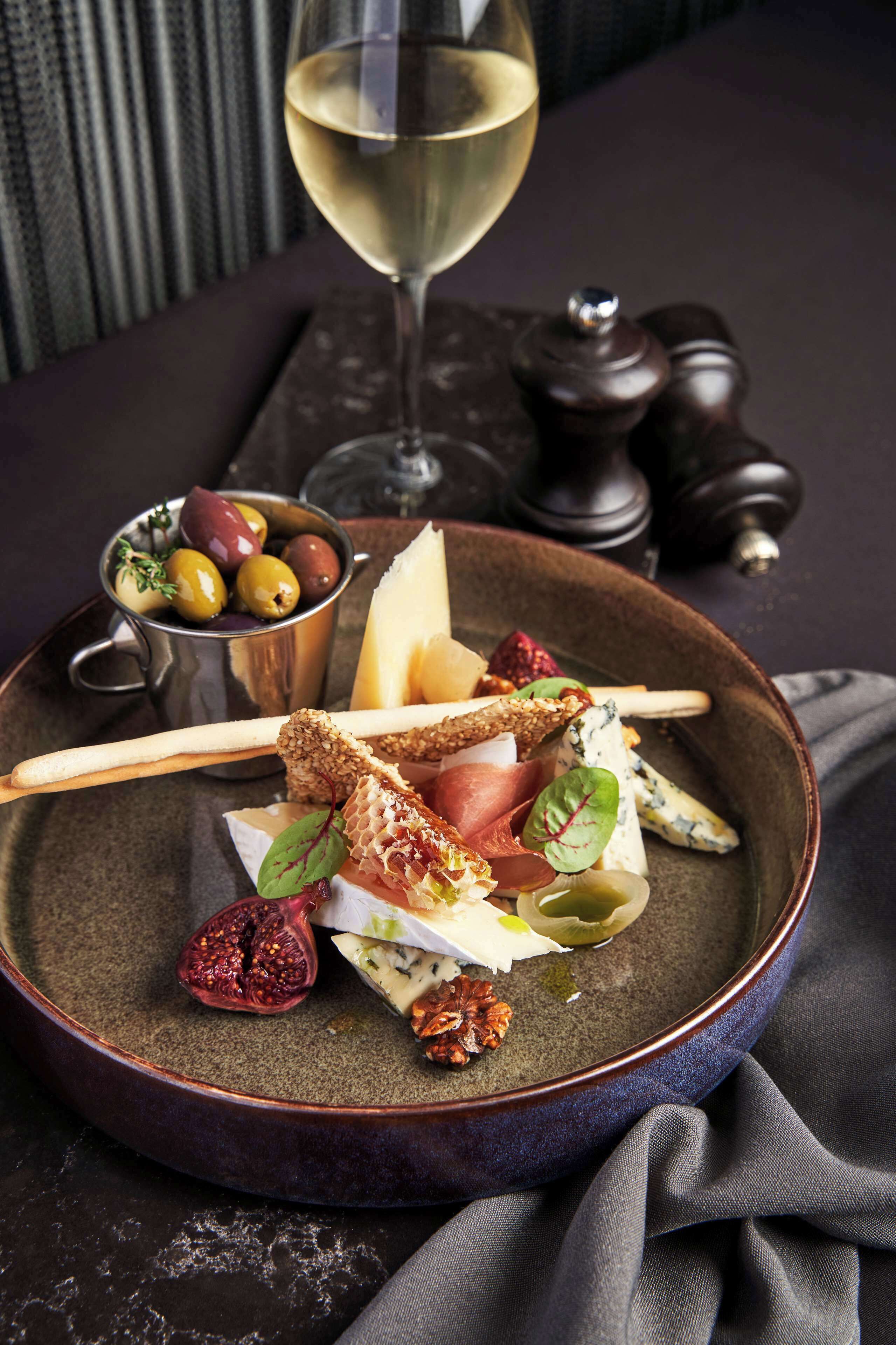 A plate of assorted cheeses, crackers, and fruits on a dark table with a glass of white wine and a salt and pepper shaker in the background.