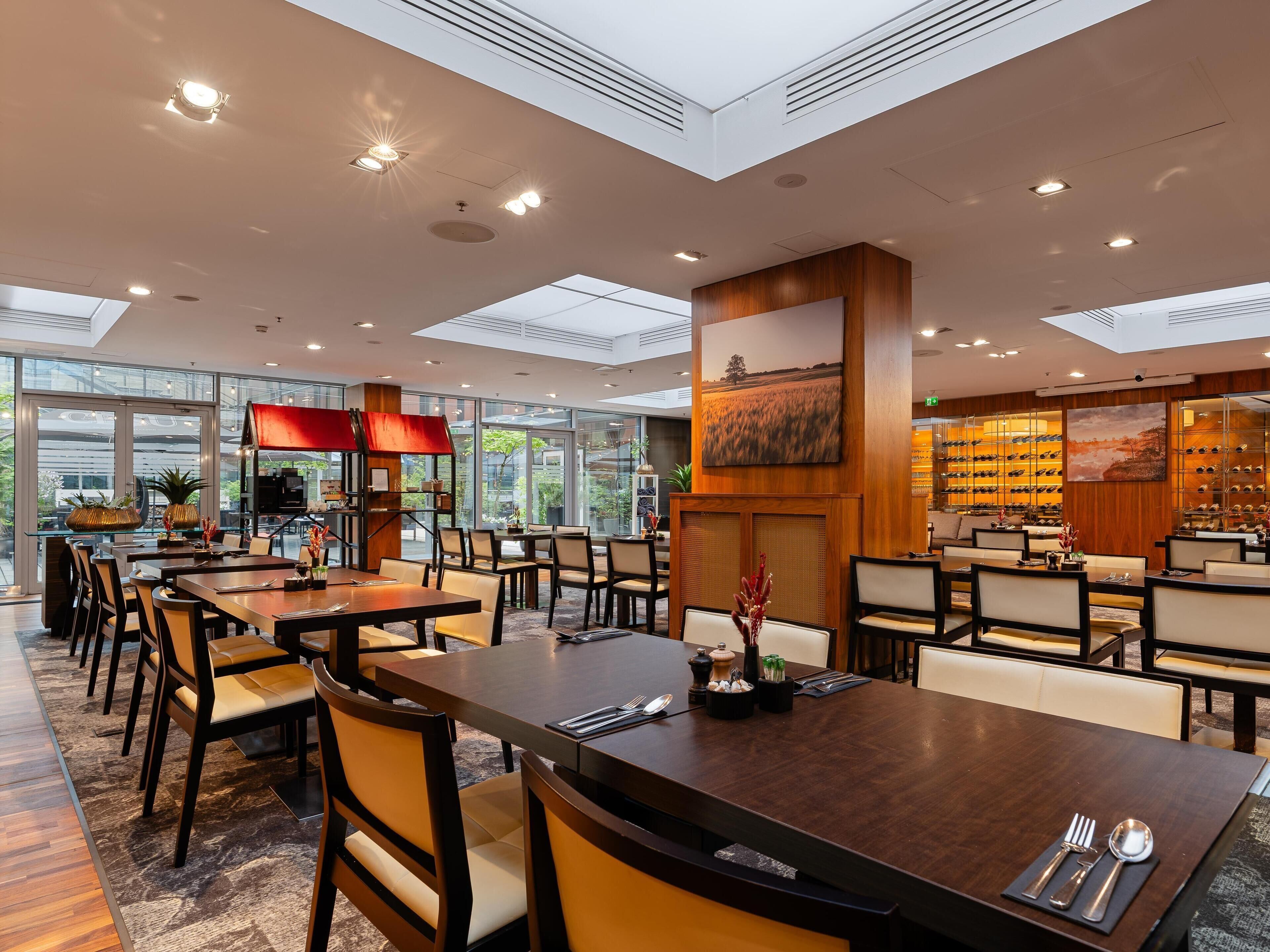 A modern hotel dining area with multiple tables and chairs, a wine rack, and large windows allowing natural light.