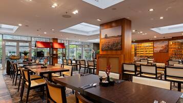 A modern hotel dining area with multiple tables and chairs, a wine rack, and large windows allowing natural light.