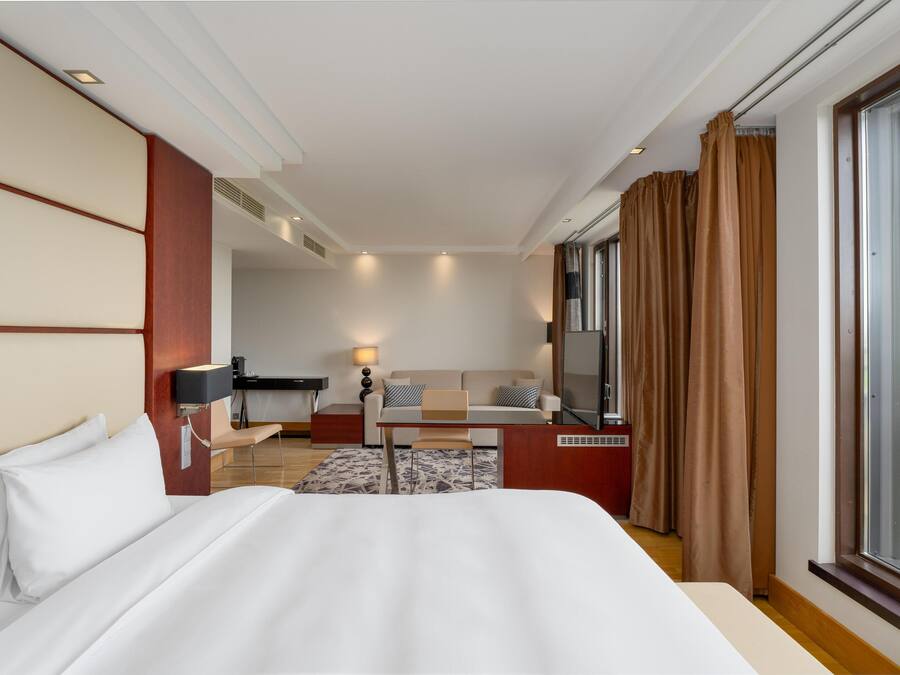 A hotel room with a large bed, a desk with a chair, a sofa, a TV, and a window with curtains.