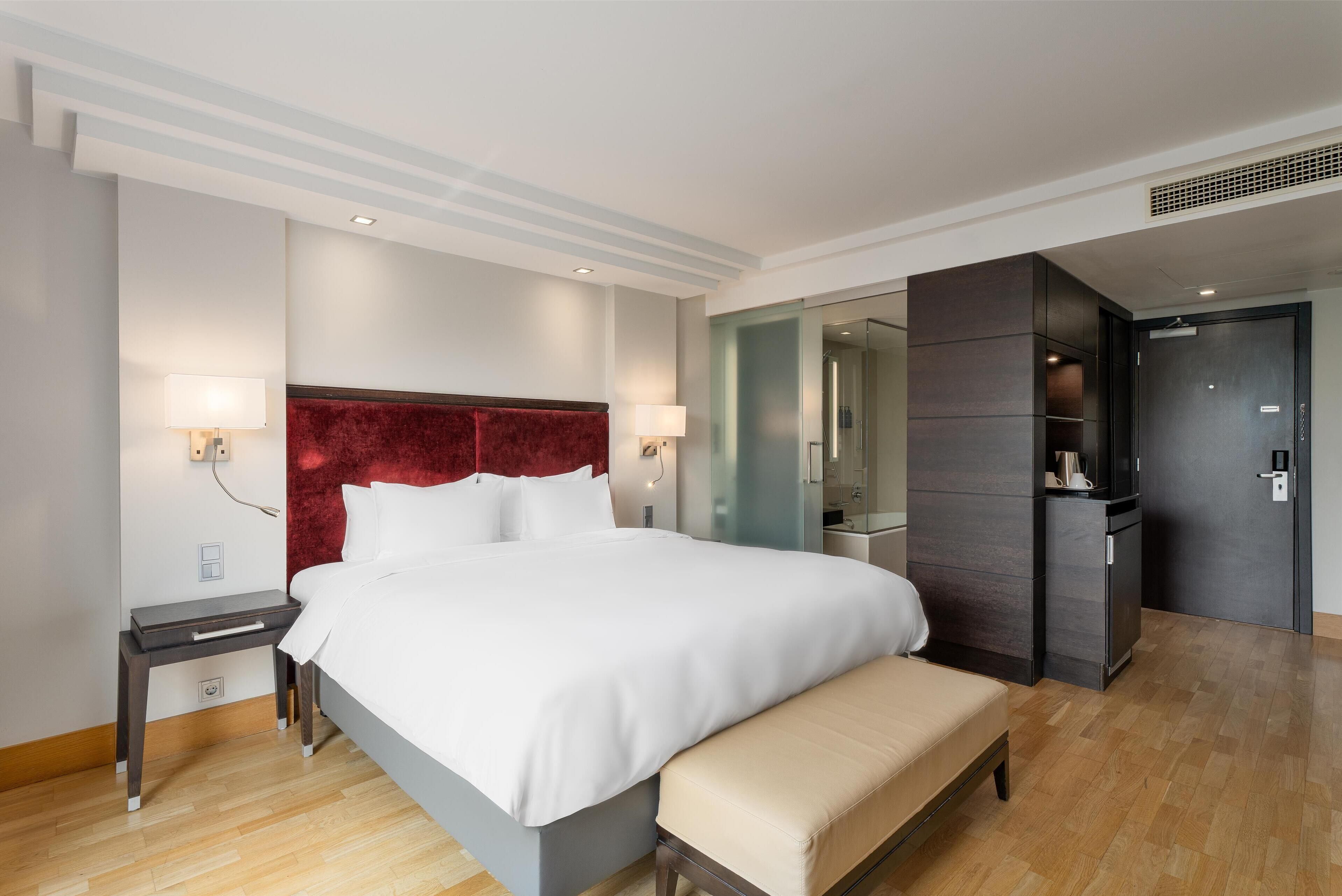 Quarto de hotel com uma cama grande, mesas de cabeceira, um banco, guarda-roupa e espelho.