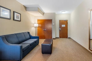 A hotel lobby with a blue sofa, a small ottoman, a floor lamp, and two wooden doors. - Comfort Inn & Suites Charleston Airport Convention Center (North Charleston)