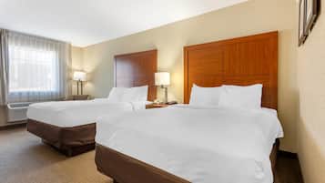 Quarto de hotel com duas camas, uma janela grande com cortinas, uma luminária fixada na parede e uma cabeceira de madeira.