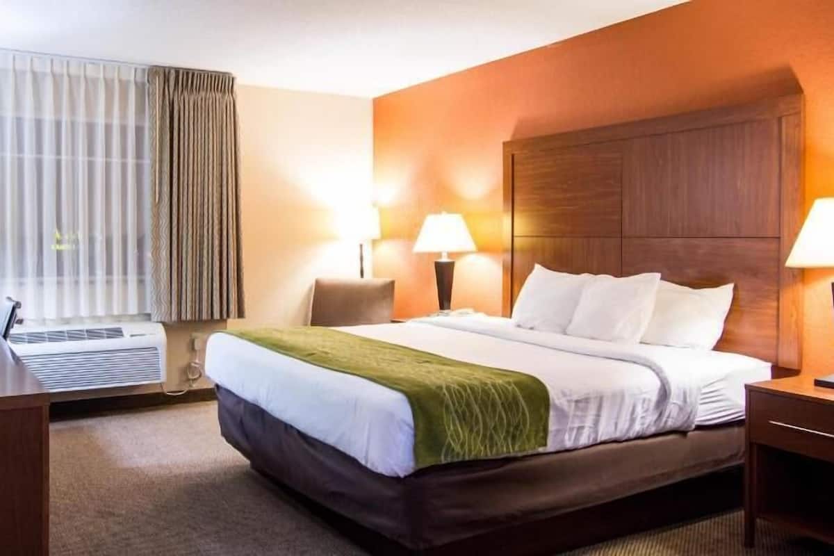 Ein Hotelzimmer mit Bett, zwei Nachttischlampen, einem Schreibtisch mit Computer, einem Fernseher und einem Fenster mit Vorhängen.