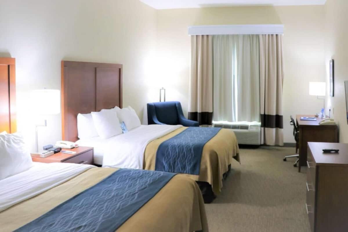 A hotel room with two beds, a desk, a chair, a lamp, and a window with curtains.