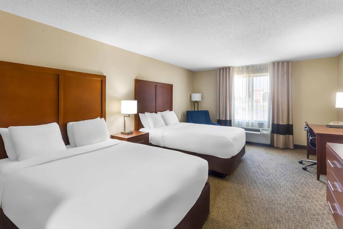 A hotel room with two beds, a desk, a chair, a lamp, and a window with curtains.