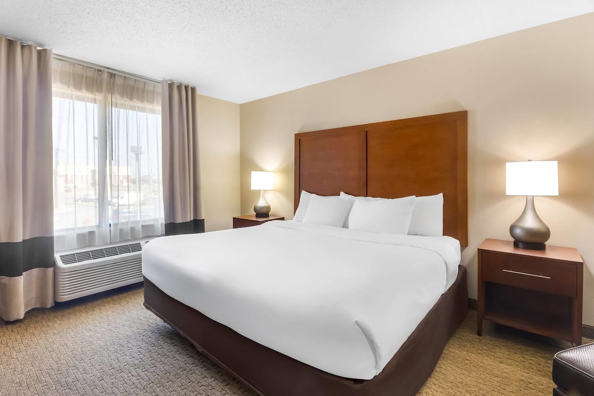 A hotel room with a large bed, two bedside tables with lamps, a window with curtains, and a wall-mounted heater.