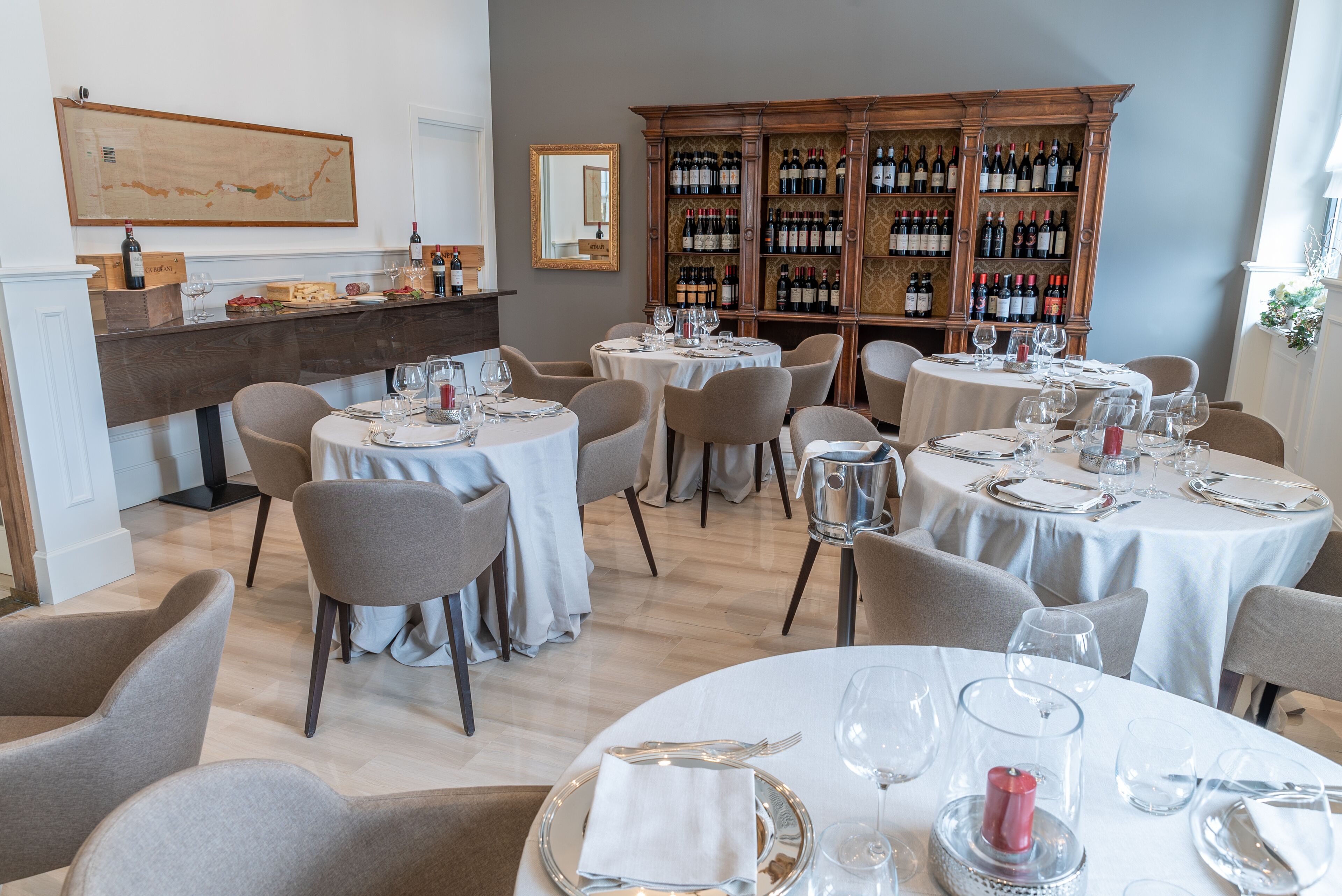 Ein Essbereich mit runden Tischen für vier Personen, jeder Tisch mit weißer Tischdecke, Stühlen und Weingläsern. Im Hintergrund ist eine Weinauslage zu sehen.