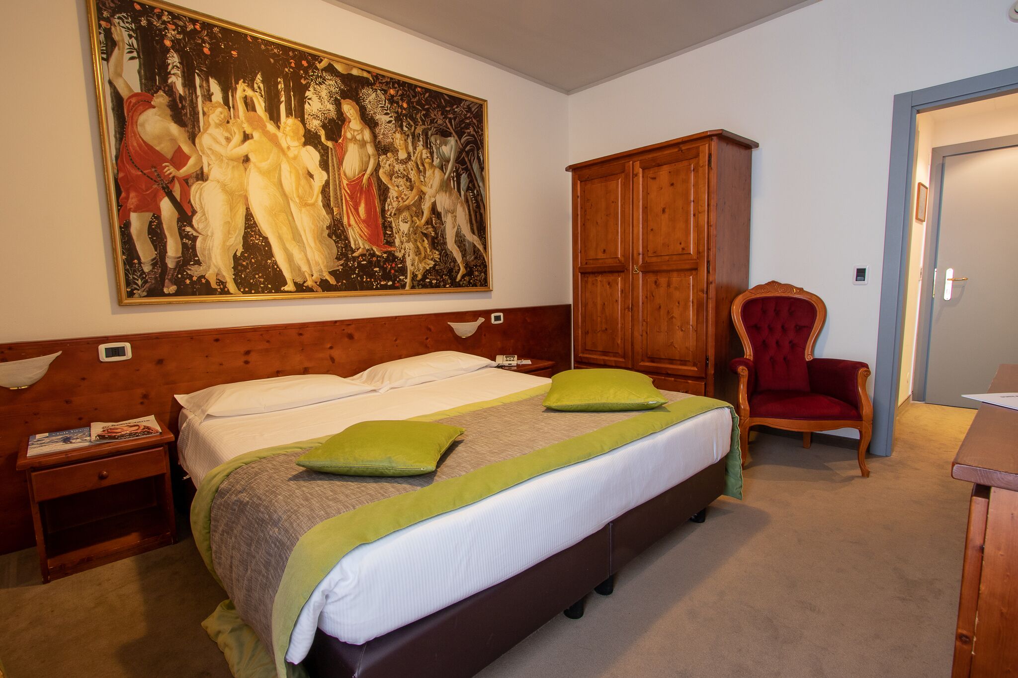 A hotel room with a large bed, a wooden wardrobe, a red armchair, and a painting on the wall.