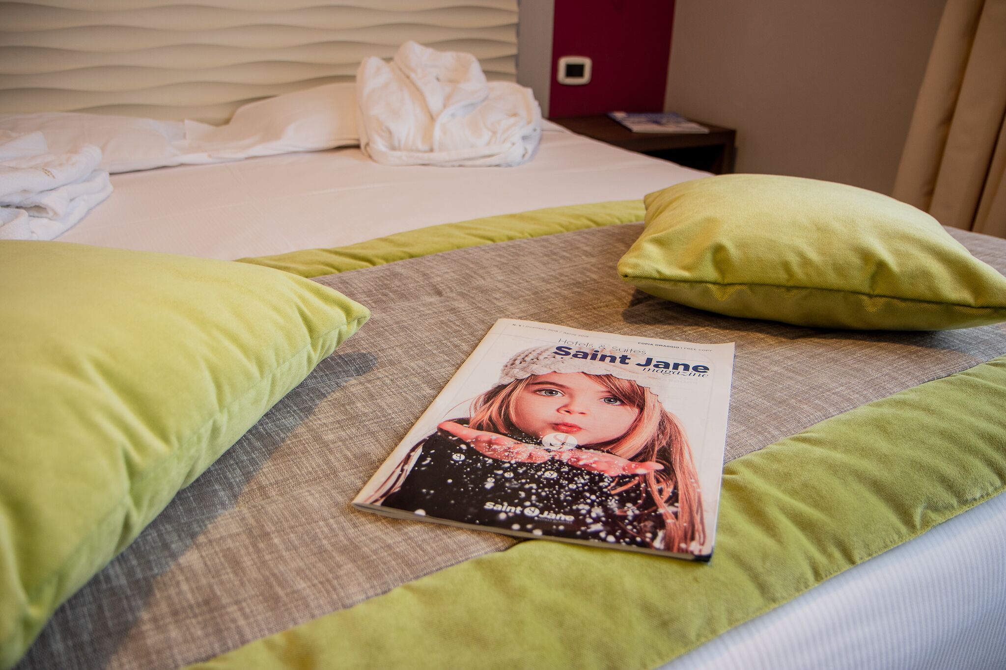 Ein Bett mit grün-grauem Bettzeug, eine Zeitschrift mit dem Titel 'Saint Jane' und ein weißes Handtuch auf dem Bett.