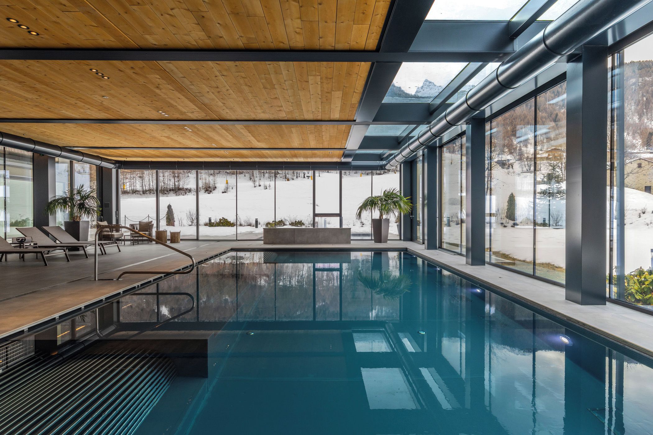 Une piscine intérieure moderne avec des parois en verre transparent, un plafond en bois et une vue sur un paysage enneigé.