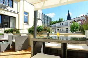 Un espacio de estar moderno al aire libre con mobiliario de mimbre y una mesa de vidrio, con vistas a un paisaje urbano con edificios históricos.