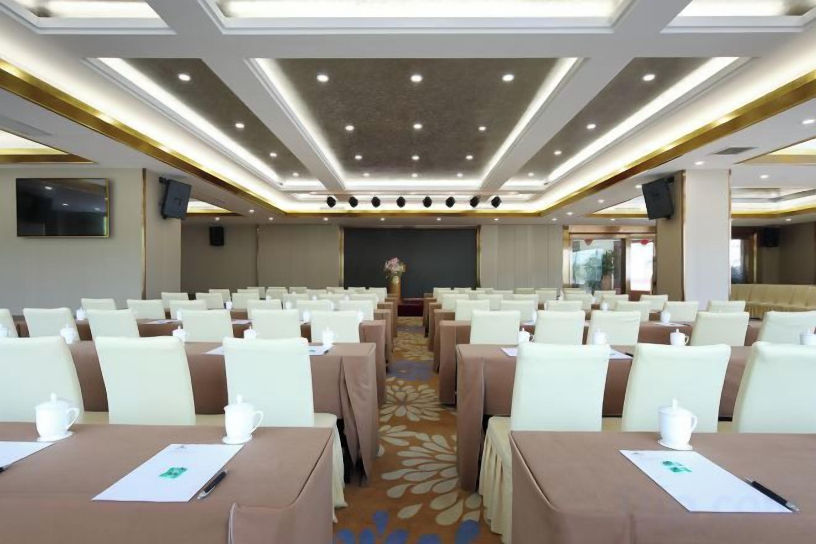 A conference room with rows of tables and chairs, a stage at the front, and a carpeted floor.