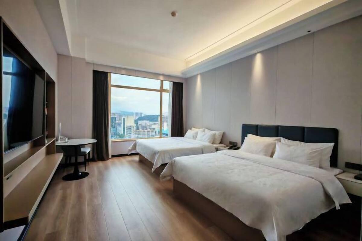 Habitación de hotel con dos camas, televisión, una mesita y un ventanal con vistas a la ciudad.