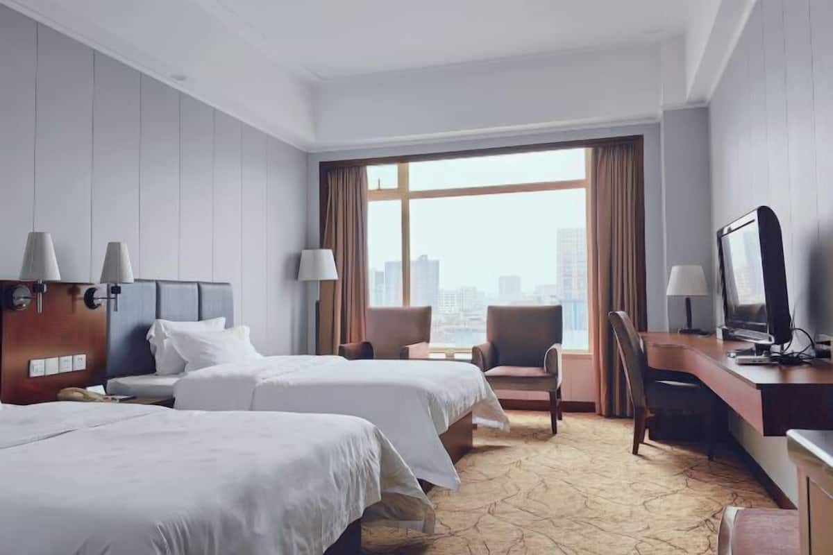 Habitación de hotel con dos camas, un escritorio, una silla, un televisor y un amplio ventanal con vistas a la ciudad.
