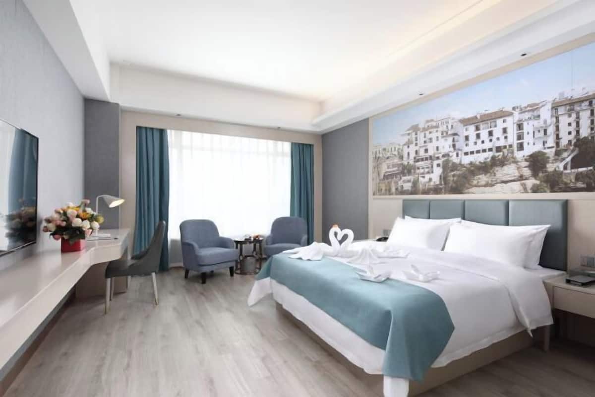 Habitación de hotel con cama, dos sillas, escritorio, televisor y un cuadro de un pueblo costero.