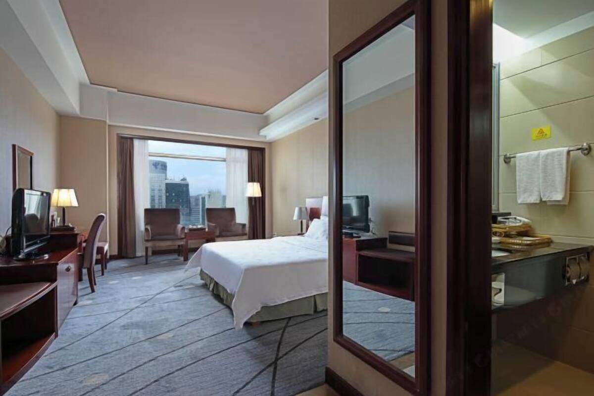 Habitación de hotel con cama, escritorio, sillas, televisor y baño con toallero.