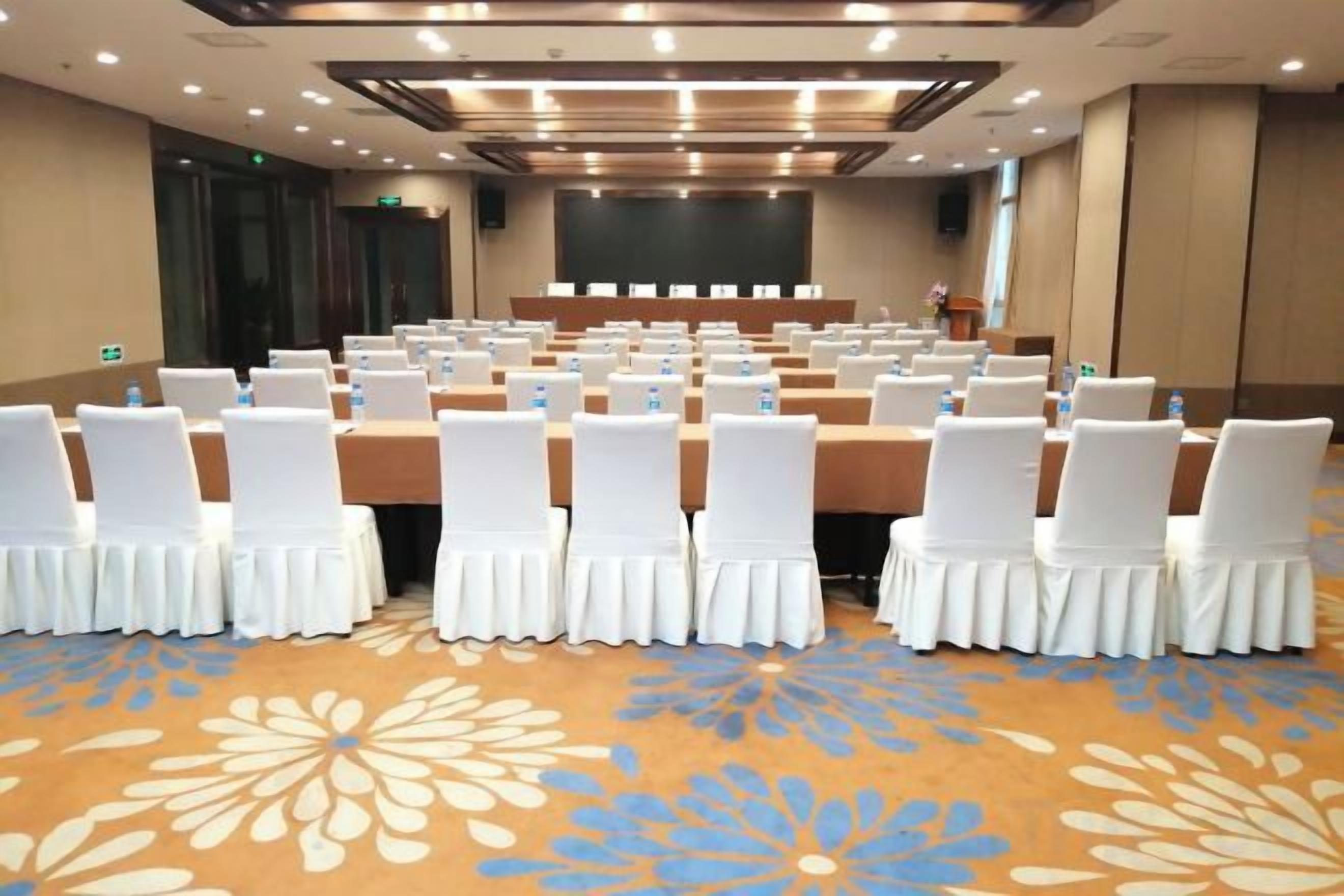 Un salón de conferencias con filas de sillas blancas, un escenario y una alfombra estampada con motivos florales.