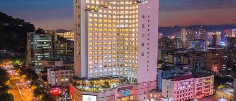 Un hotel de varios pisos con una torre central destacada, rodeado de otros edificios, y una calle concurrida con vehículos y letreros iluminados.