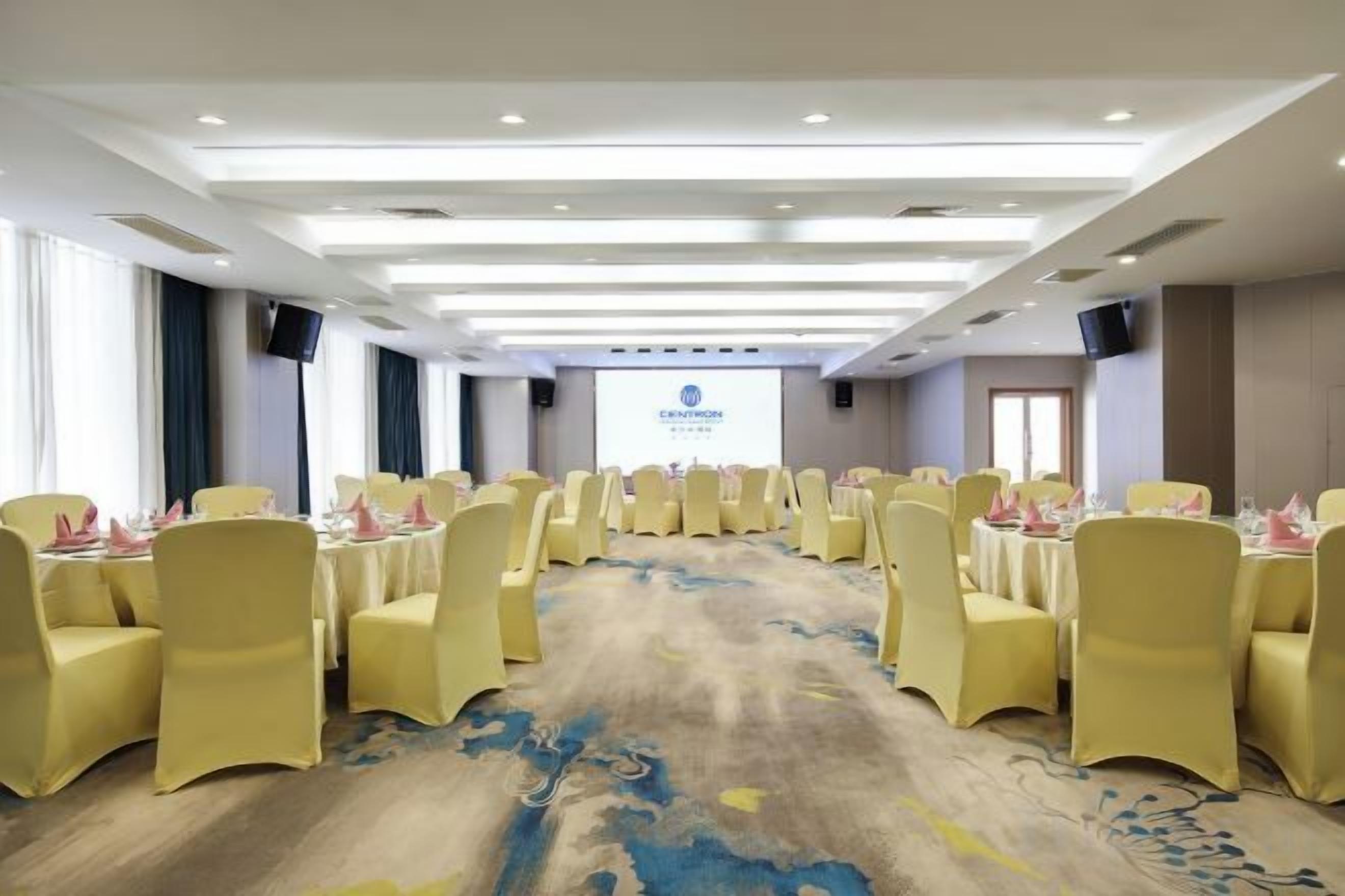 Un salón de conferencias con mesas y sillas redondas, piso alfombrado y una pantalla de proyección al frente.