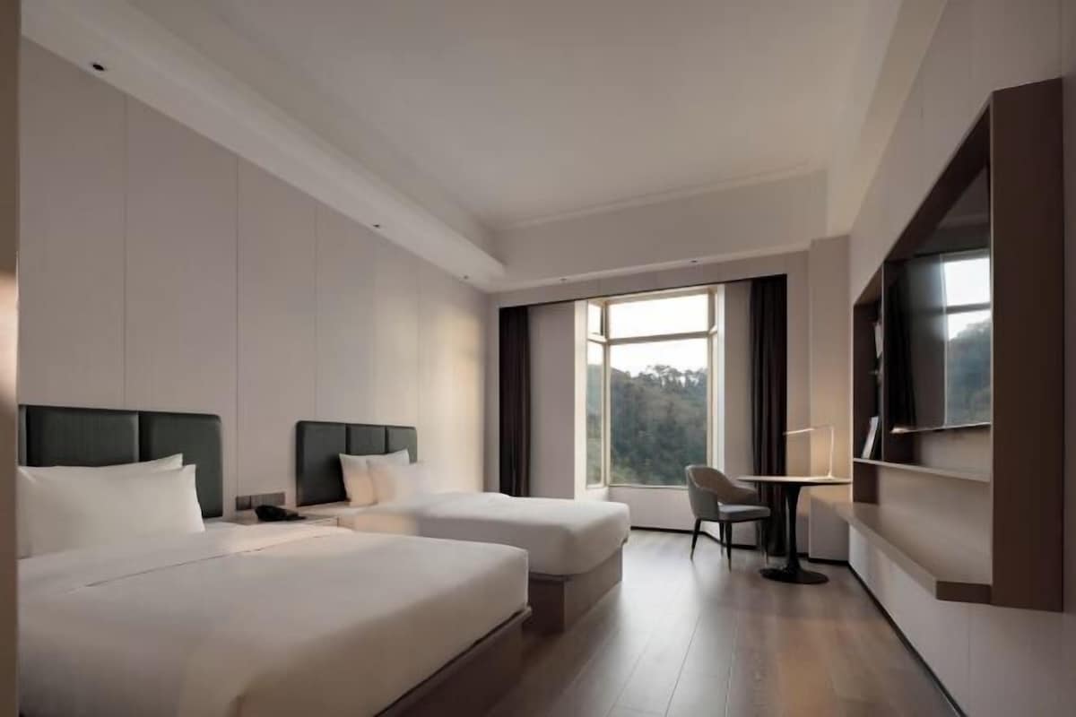 A hotel room with two beds, a desk, a chair, a TV, and a large window with a view.