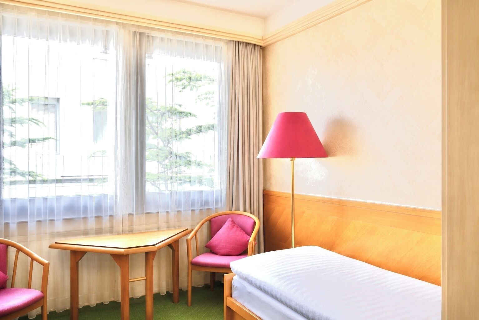 Une chambre d’hôtel avec un lit, un bureau, deux chaises, une lampe et une fenêtre avec des rideaux.