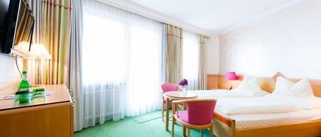 Una habitación de hotel con cama, escritorio, silla, televisión y ventana con cortinas.