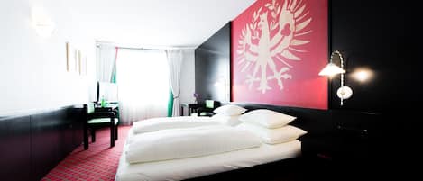 Una habitación de hotel con una cama grande, un escritorio y una silla. Una pared roja y negra con un emblema blanco.