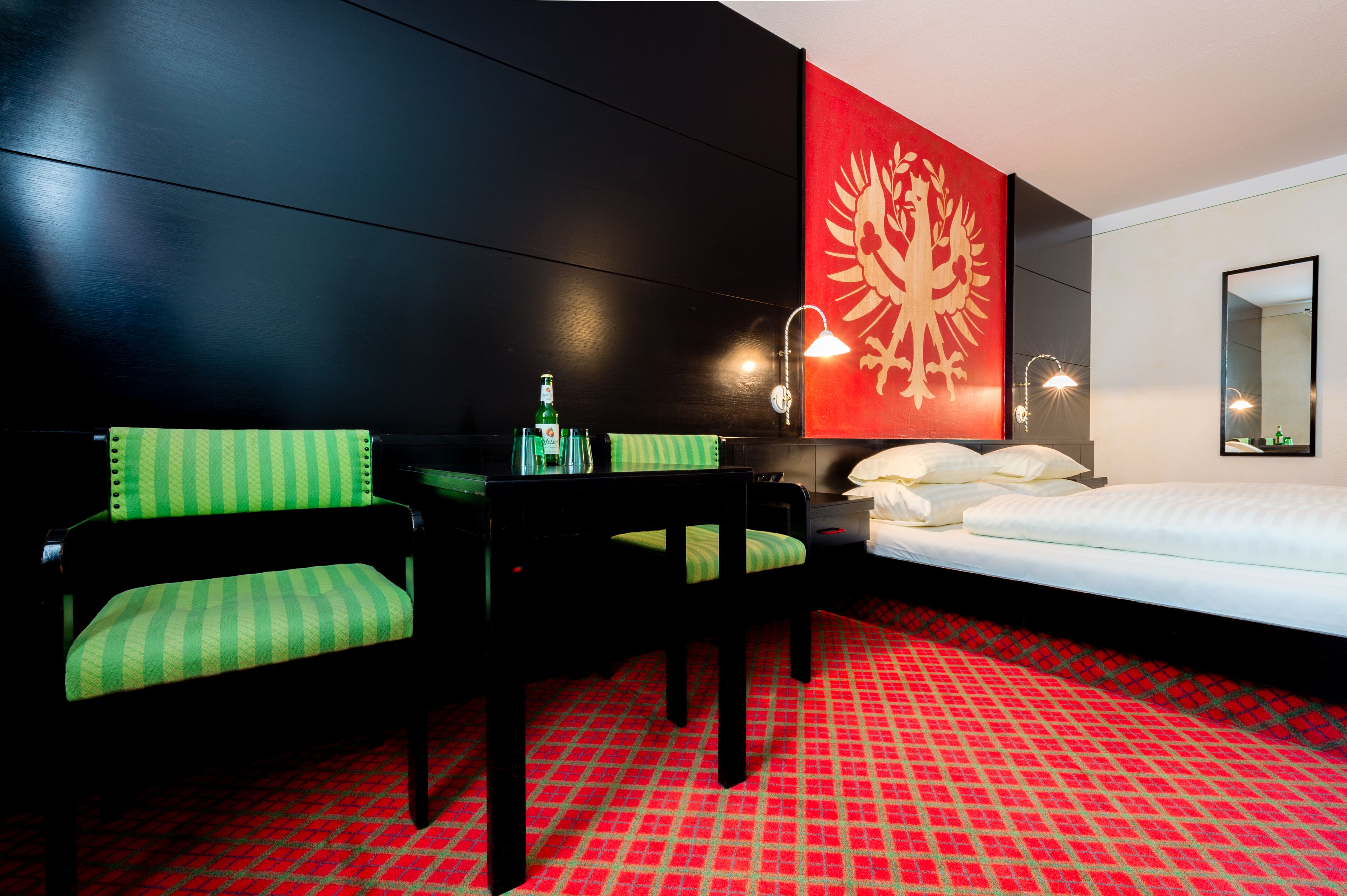 Habitación de hotel con una pared negra, alfombra estampada en rojo y blanco, una cama, una silla verde y negra, una mesita con una botella y una lámpara de pared.