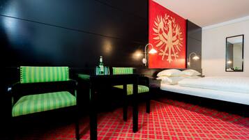 Camera d'albergo con parete nera, tappeto rosso e bianco, un letto, una sedia verde e nera, un tavolino con una bottiglia e una lampada a parete.