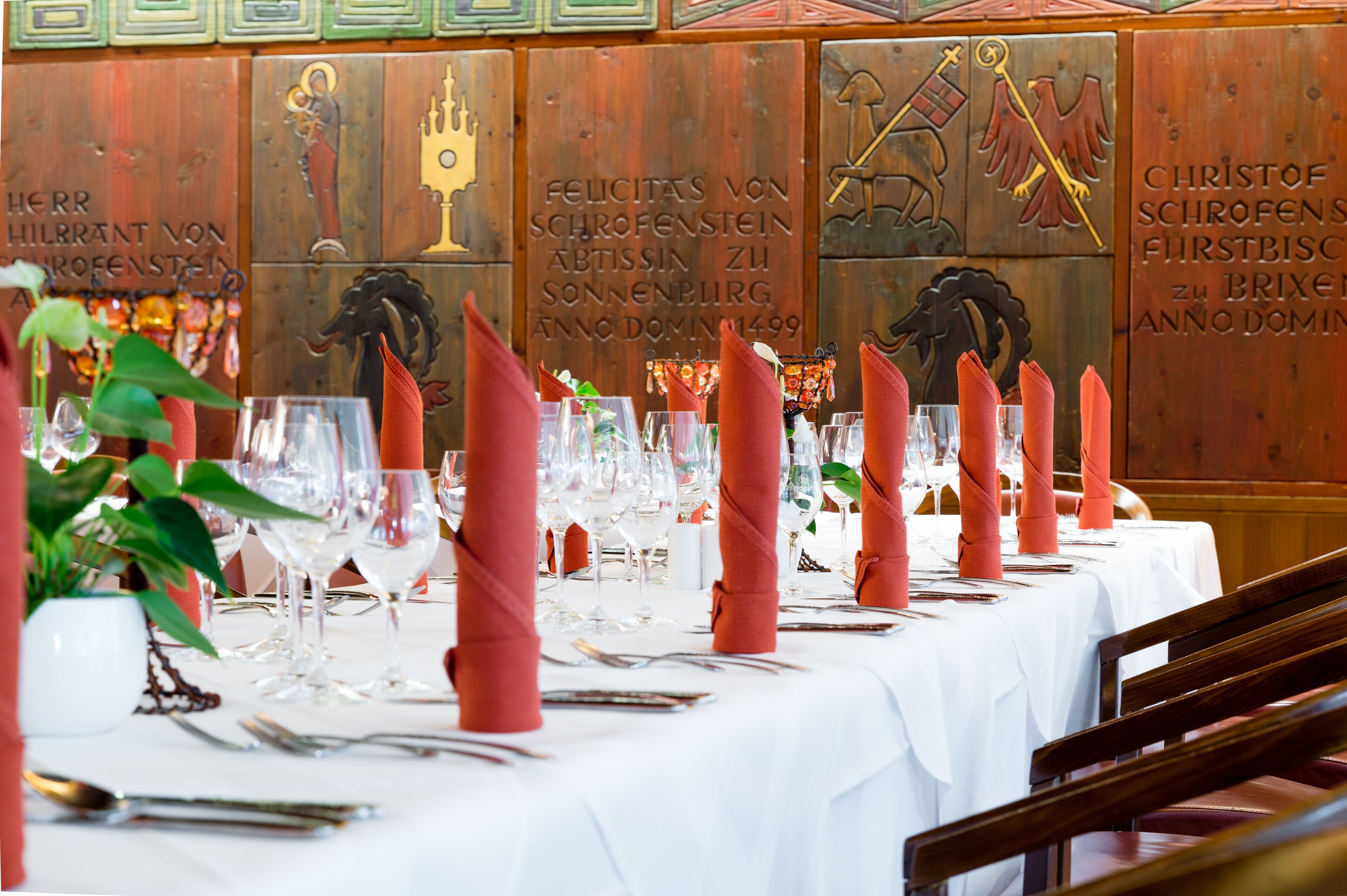 A restaurant setting with a long table set for a meal, featuring neatly folded red napkins, wine glasses, and cutlery.