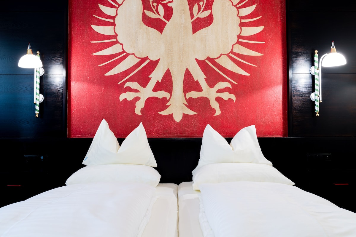 Uma cama com lençóis e almofadas brancos, uma parede vermelha com brasão branco, e duas luminárias fixadas na parede.