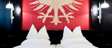 A bed with white linens and pillows, a red wall with a white coat of arms, and two wall-mounted light fixtures.