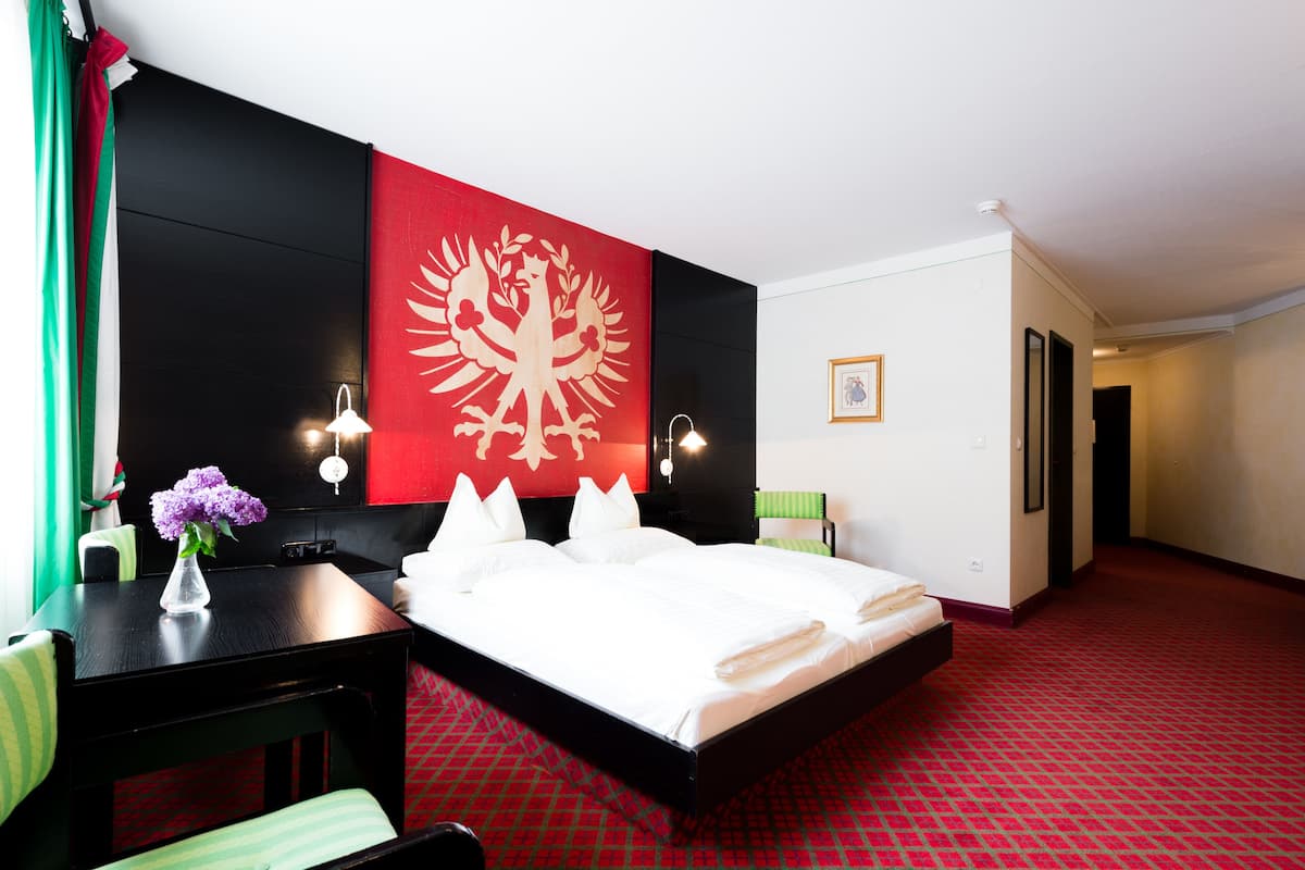 Quarto de hotel com uma cama, uma mesa, uma cadeira, um vaso com flores e uma parede preta e vermelha com um brasão.