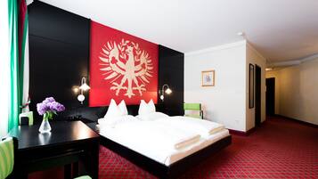 Une chambre d’hôtel comprenant un lit, un bureau, une chaise, un vase avec des fleurs et un mur rouge et noir orné d’un blason.